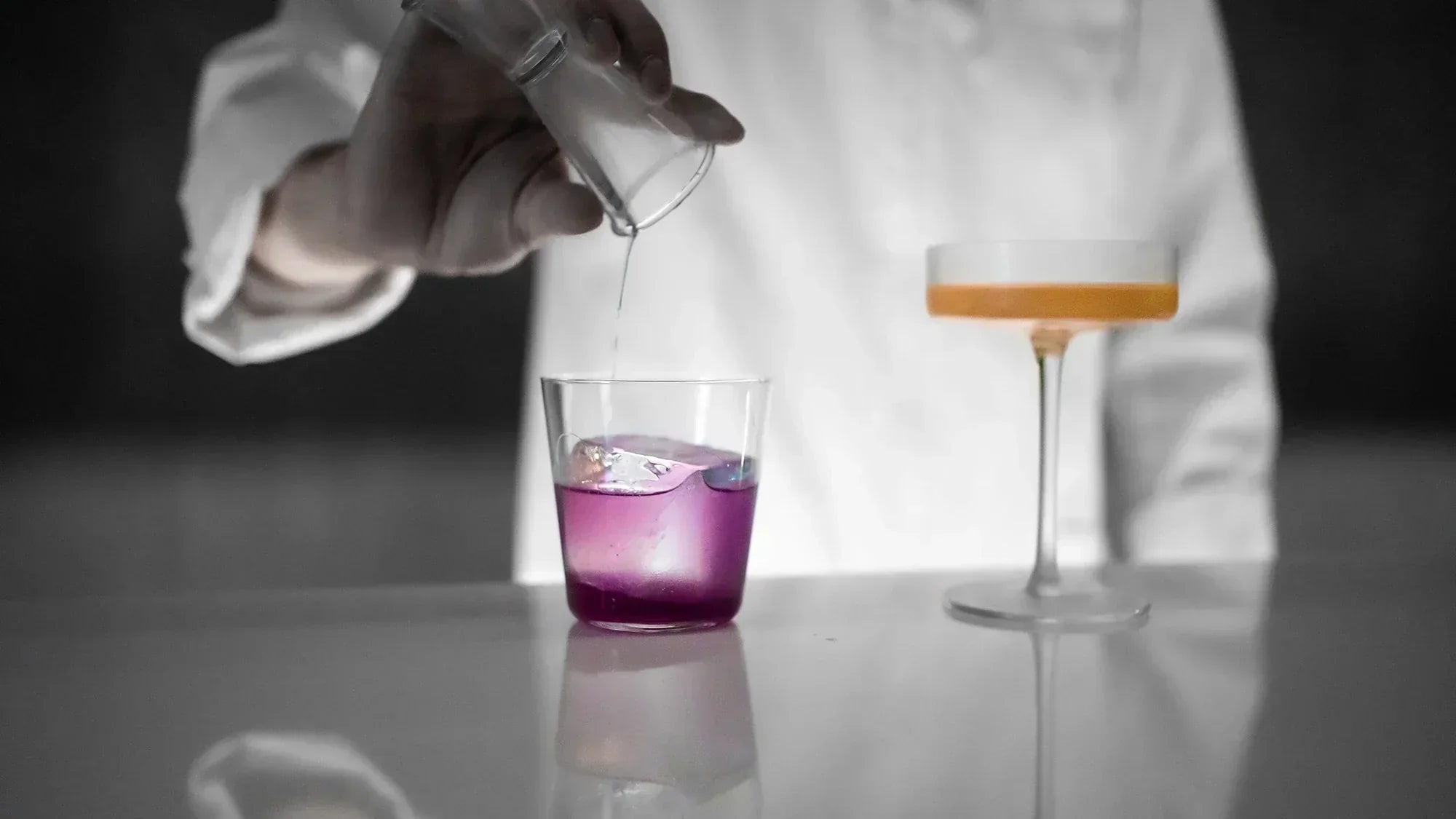 Scientist pouring clear liquid into a glass with purple solution beside an orange liquid in a lab setting