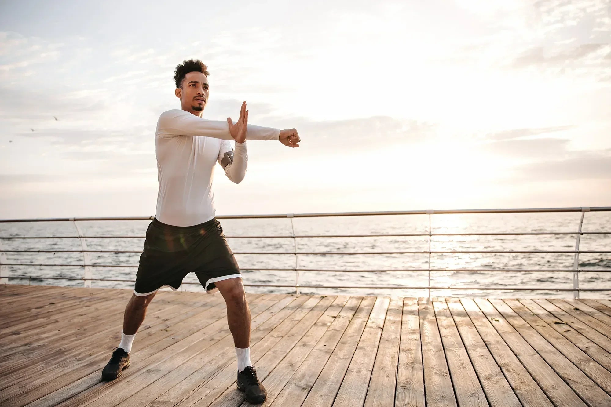 Athletic man in sportswear stretching on wooden pier by the sea at sunrise