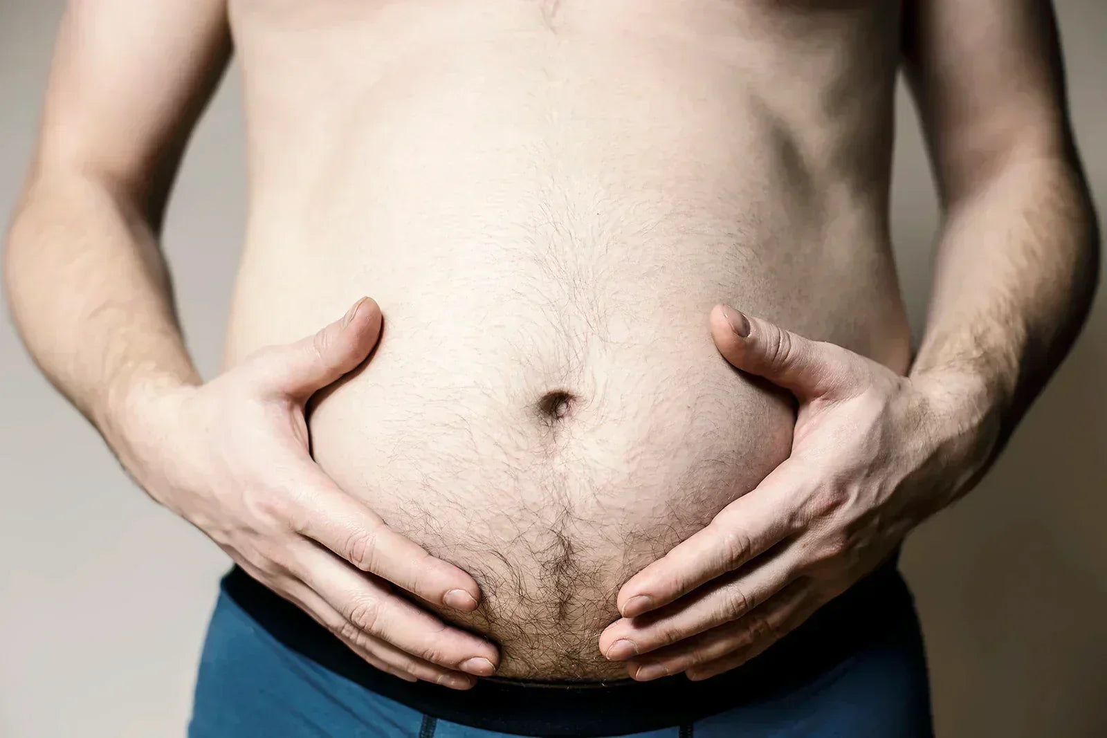 Close-up of a man's bare stomach with visible body fat, hands resting on belly