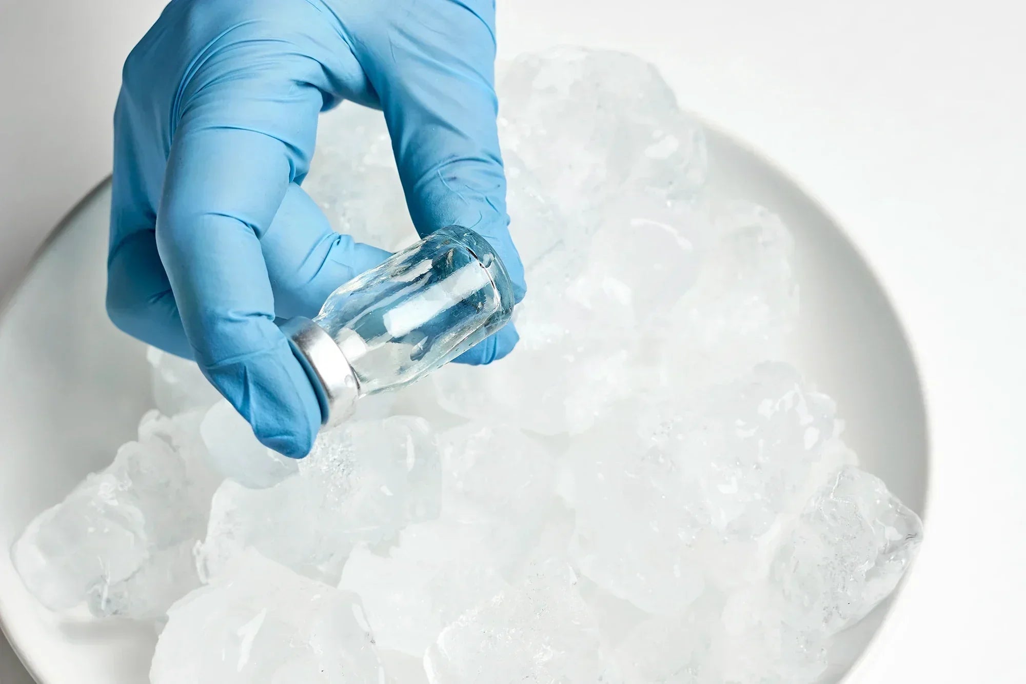 Gloved hand holding a vaccine vial from a bowl filled with ice cubes on a white background