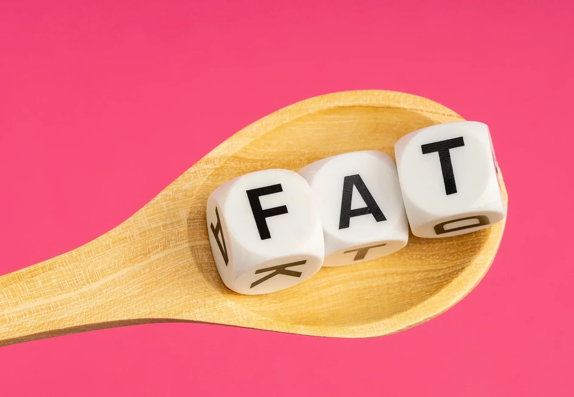 Wooden spoon holding dice spelling 'FAT' on pink background, body fat health concept